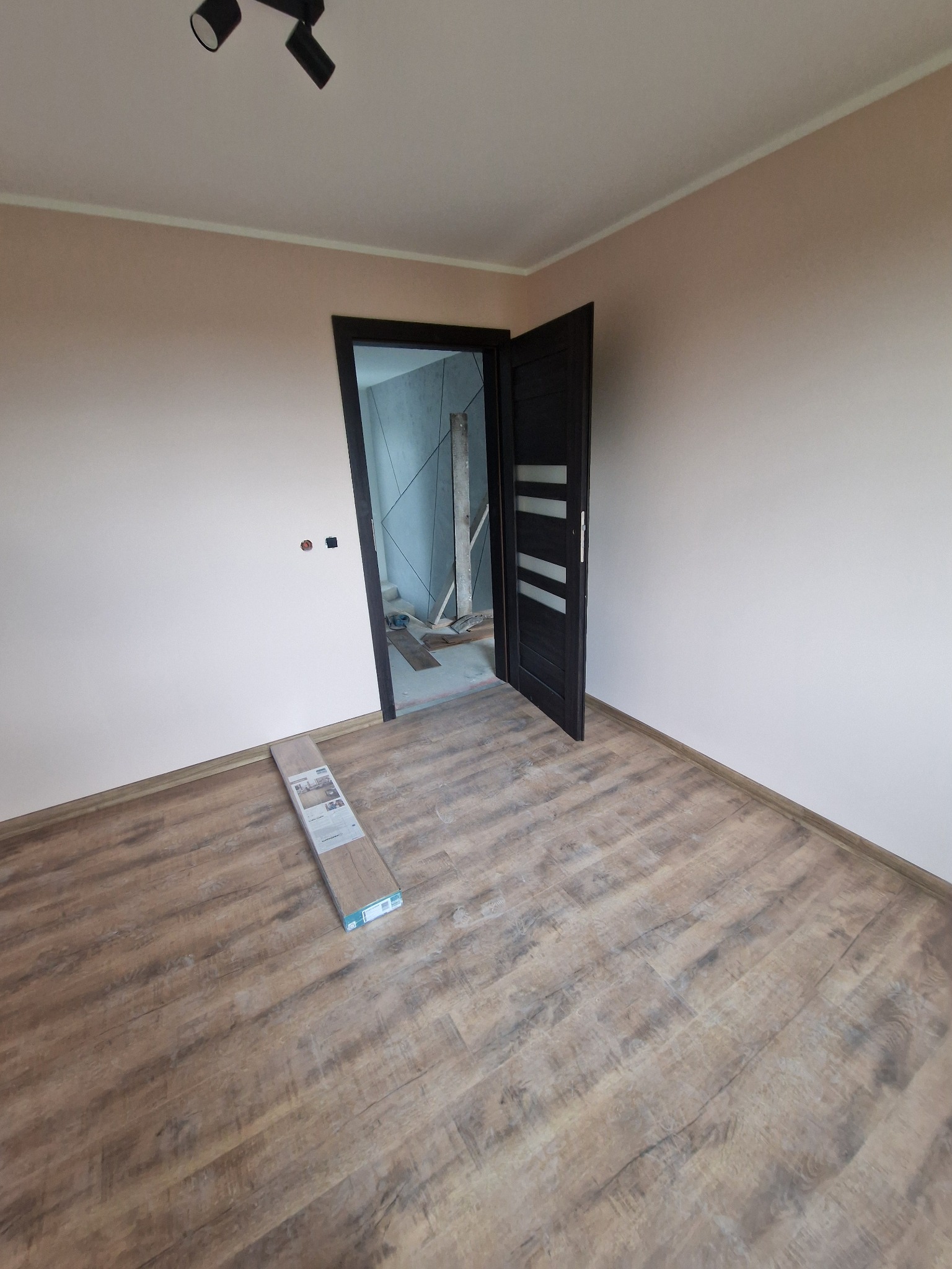 Pokój po remoncie z drewnianą podłogą i otwartymi, ciemnymi drzwiami. Na podłodze leżą panele podłogowe. Widoczne gniazdka elektryczne na ścianie.