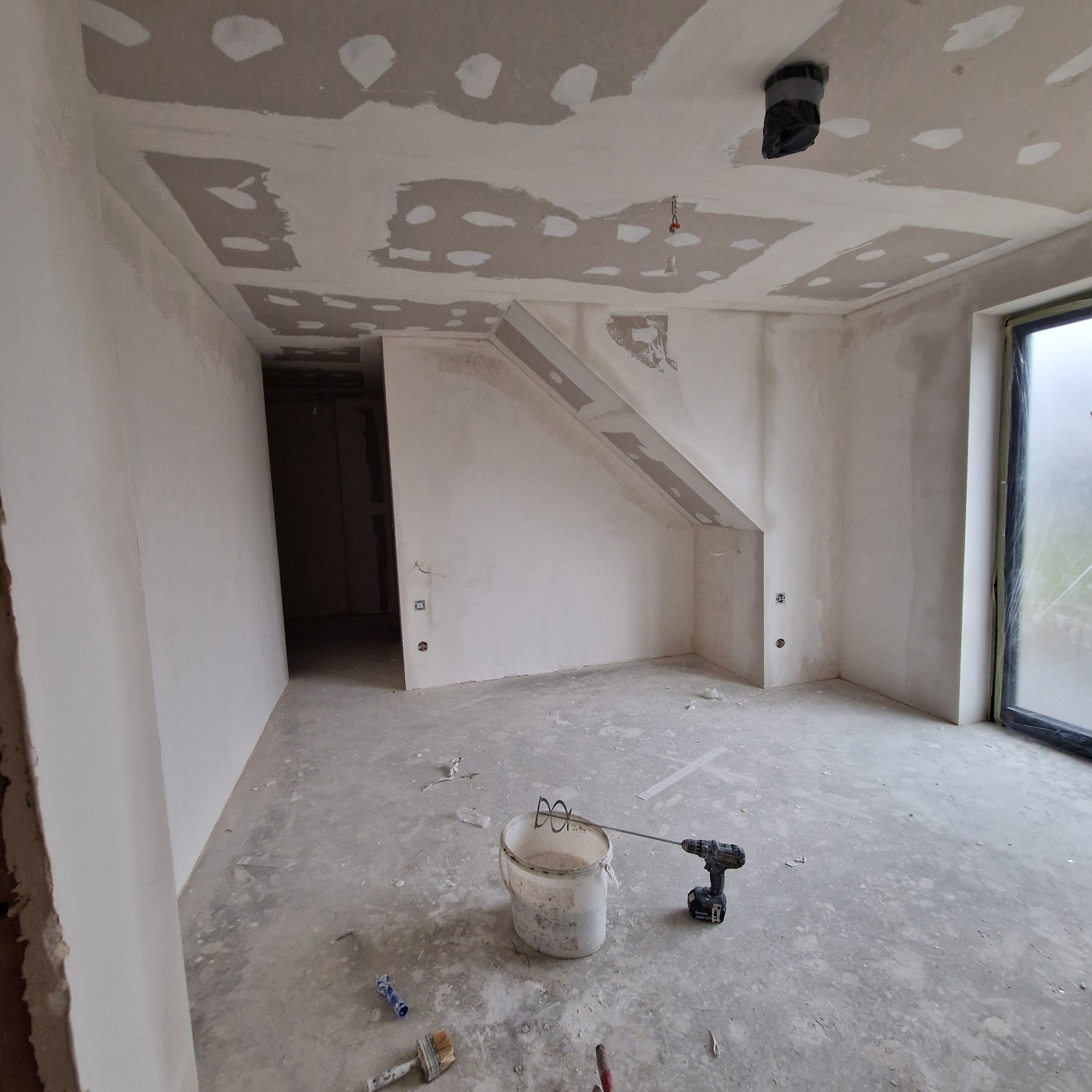 Wnętrze w trakcie prac wykończeniowych: surowe ściany z widocznymi łatami gipsowymi, narzędzia malarskie i wiadro z mieszadłem na podłodze. Okno z widokiem na zewnątrz.