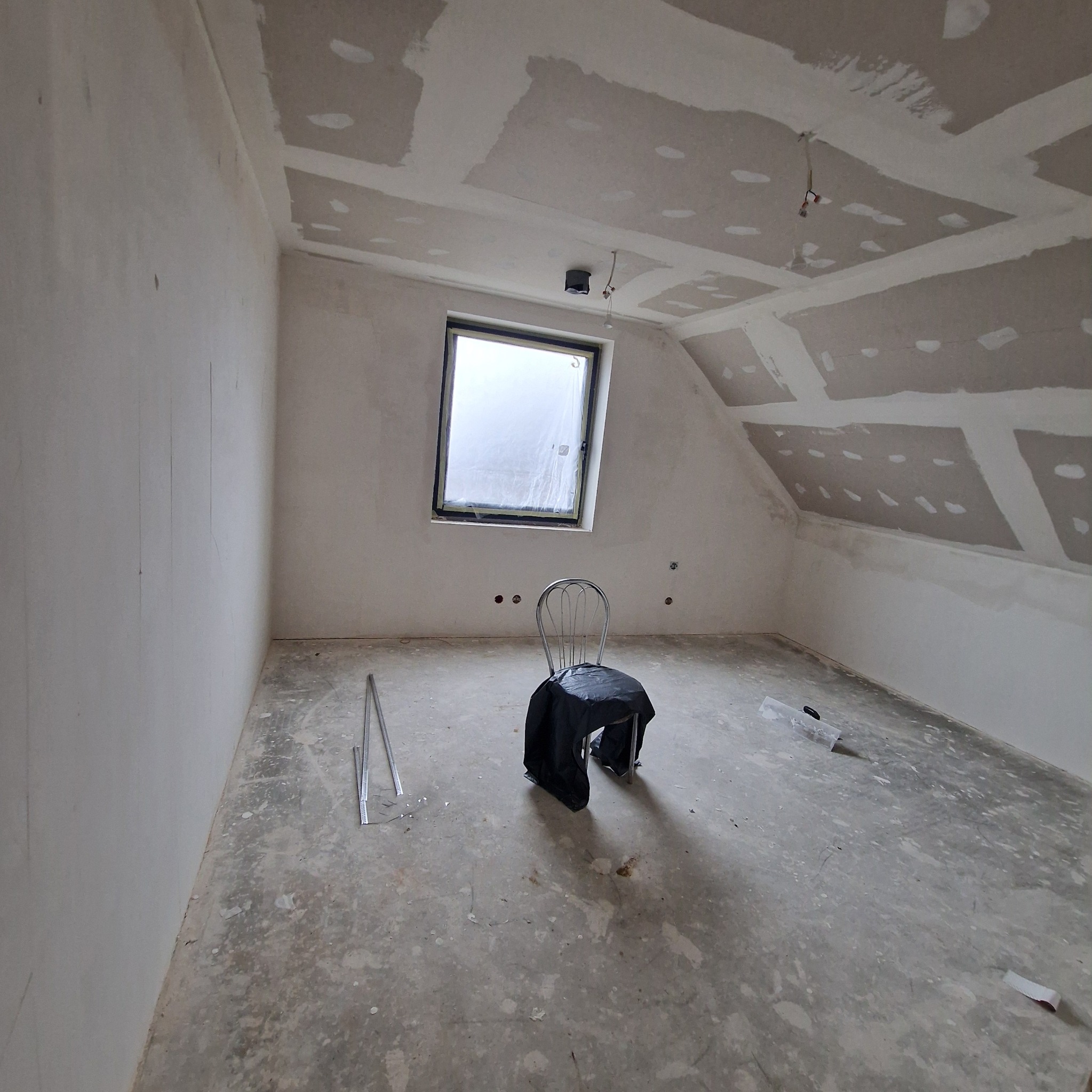 Poddasze w trakcie prac wykończeniowych: ściany i sufit z płyt gipsowo-kartonowych, okno dachowe z folią ochronną, krzesło przykryte folią. Widoczne ślady po szpachlowaniu.