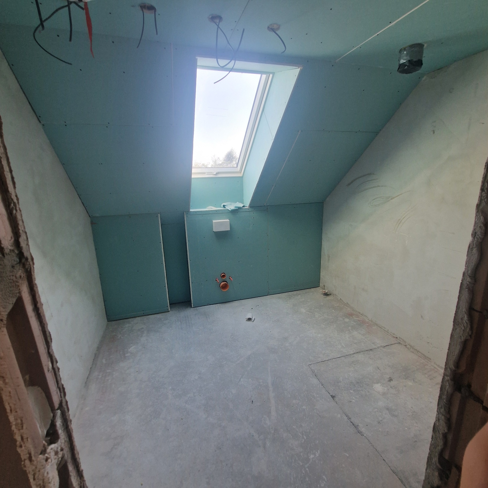 Surowe wnętrze poddasza z oknem dachowym, ściany wyłożone płytami gipsowo-kartonowymi, widoczne instalacje. Etap prac wykończeniowych w nowym budownictwie.