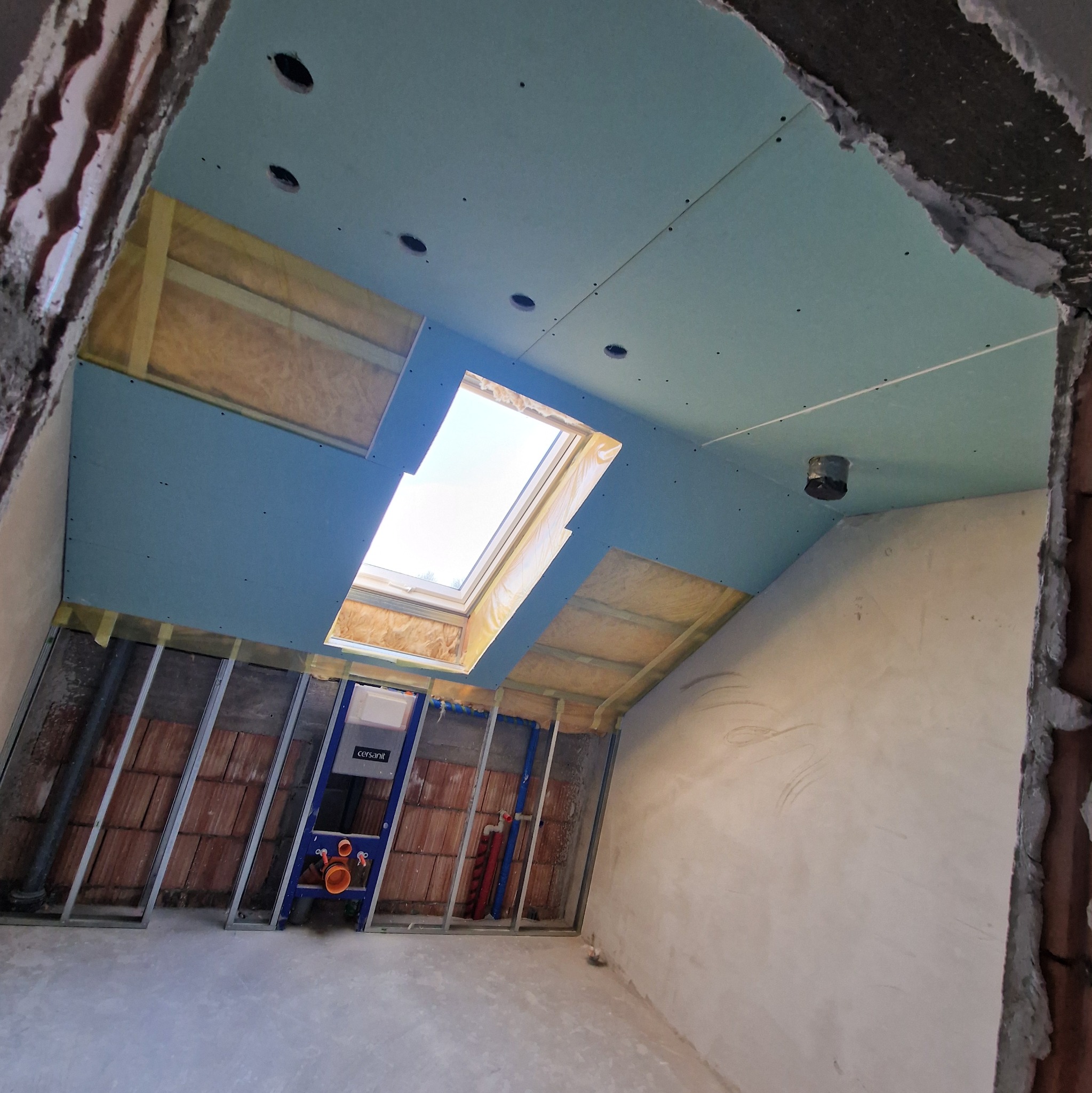 Łazienka na poddaszu w trakcie remontu: zamontowane płyty gipsowe na suficie z oknem dachowym, stelaż podtynkowy WC, instalacje. Widoczne cegły i surowy tynk.