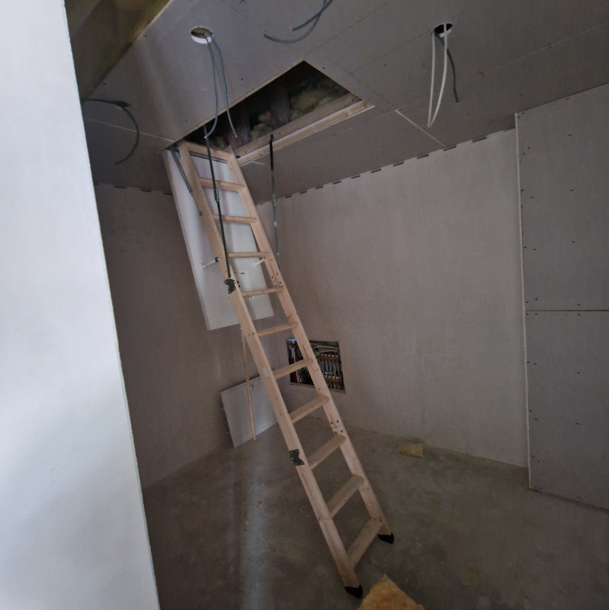 Drewniana drabina strychowa rozłożona w surowym wnętrzu. Widoczne przewody elektryczne i płyty gipsowo-kartonowe na ścianach. Etap prac wykończeniowych.
