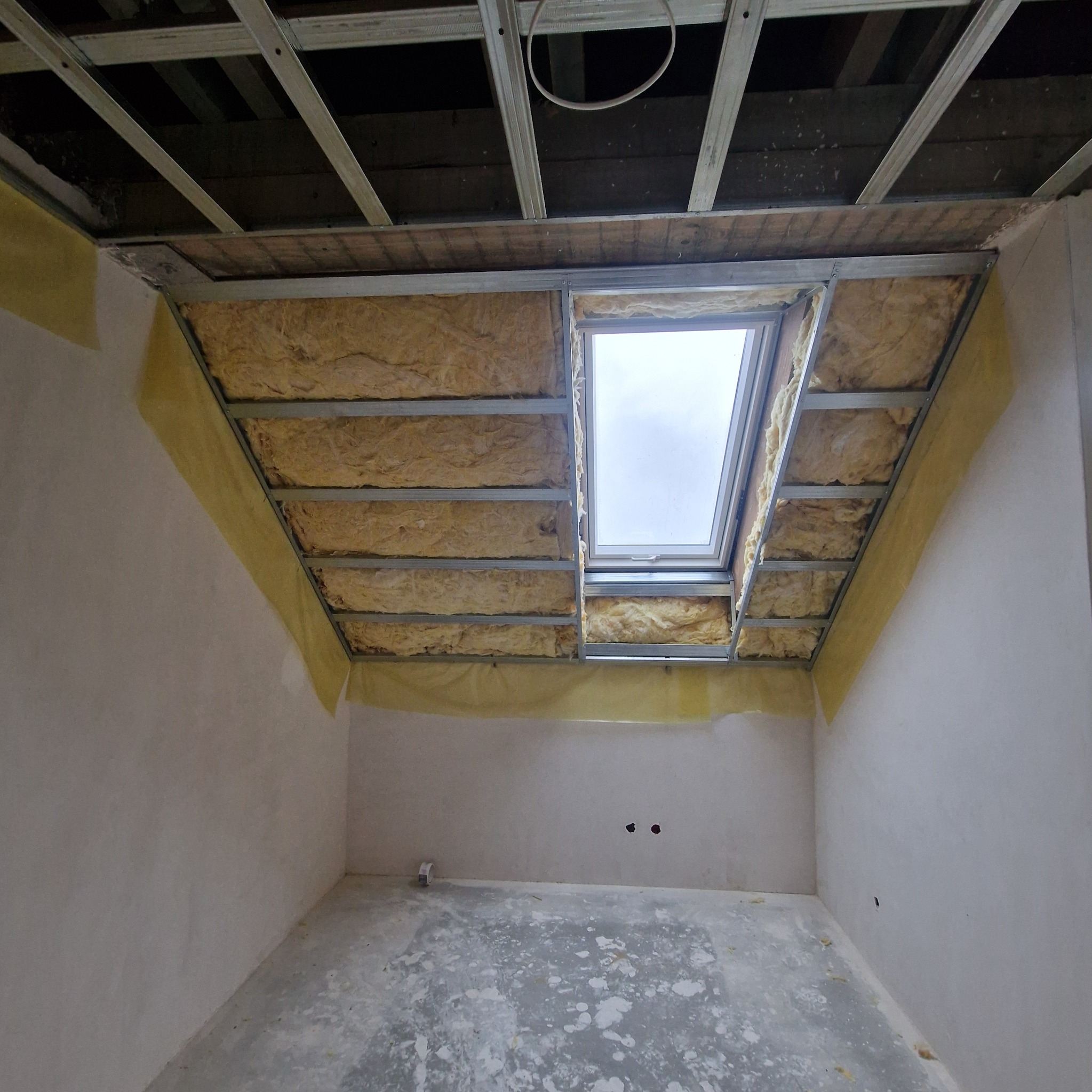 Montaż okna dachowego w surowym wnętrzu. Widoczne ocieplenie wełną mineralną i metalowa konstrukcja pod płytę gipsową. Przygotowanie do wykończenia poddasza.