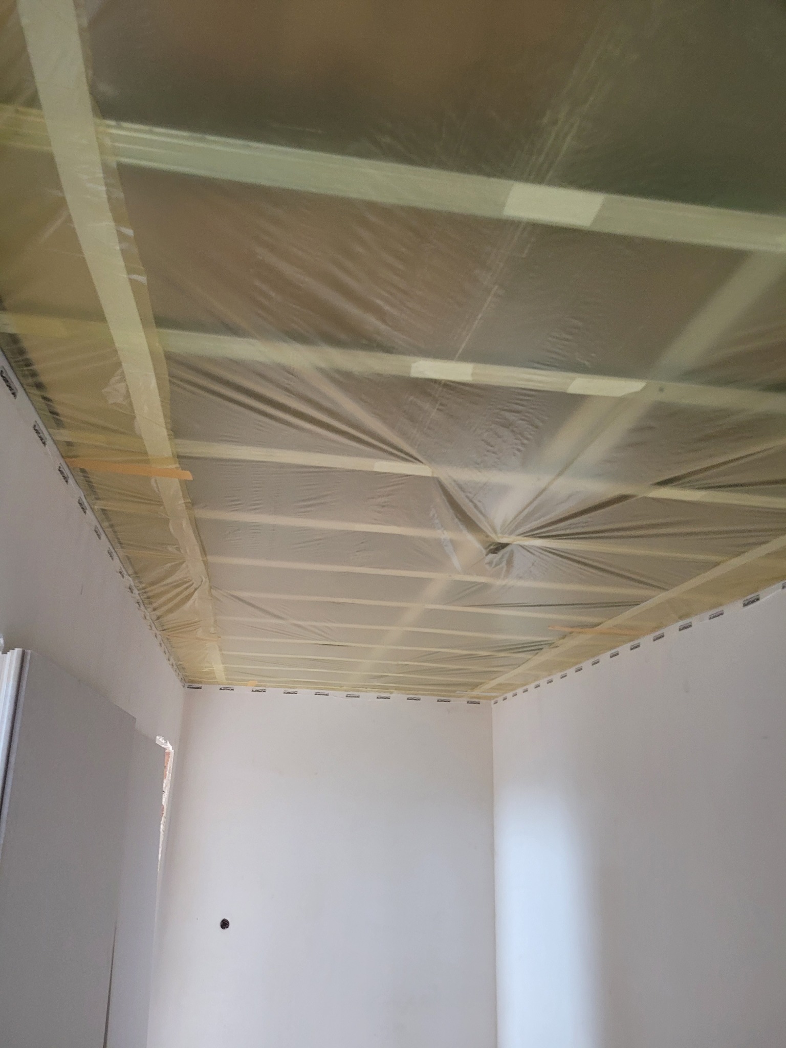 Przygotowanie sufitu do prac wykończeniowych: drewniana konstrukcja zabezpieczona folią i taśmą malarską w rogu pomieszczenia z fragmentem ściany z płyty gipsowej.