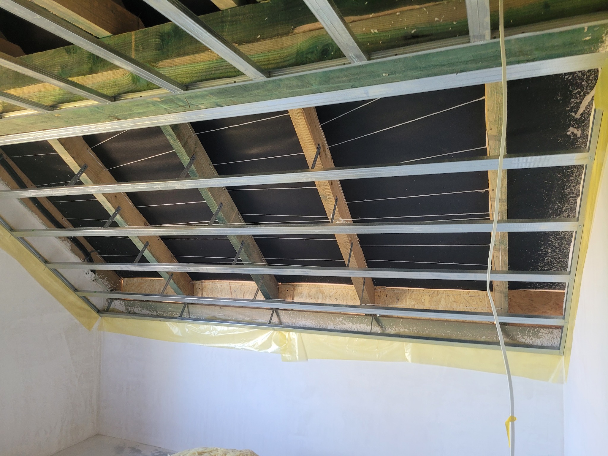 Szkielet sufitu podwieszanego z metalowych profili, widoczne drewniane elementy konstrukcyjne i folia ochronna na ścianach. Etap prac wykończeniowych w pomieszczeniu.