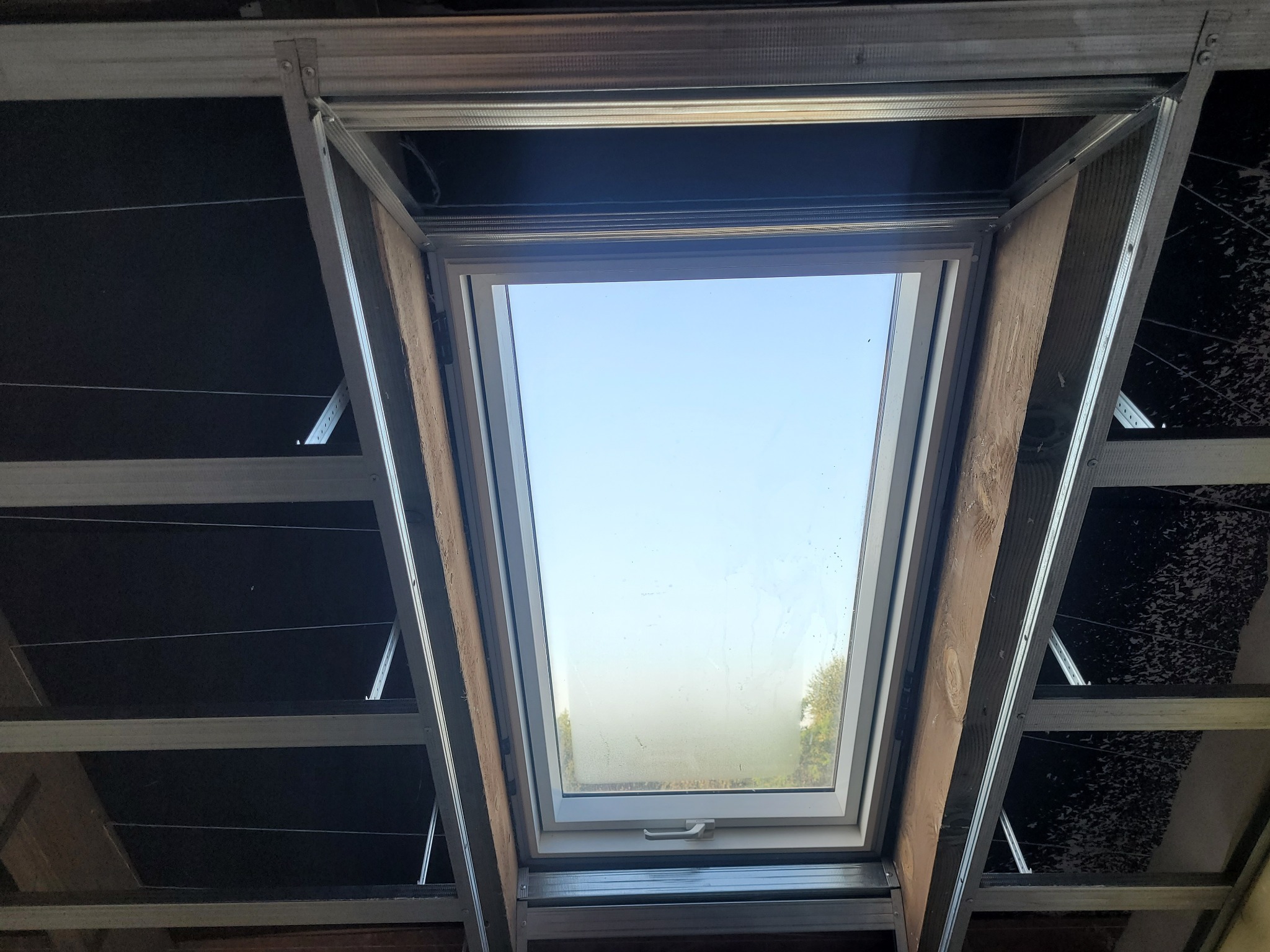 Montaż okna dachowego w konstrukcji metalowej, widok od wewnątrz. Widoczne elementy mocujące i fragment nieba przez szybę. Drewniane elementy wykończeniowe.