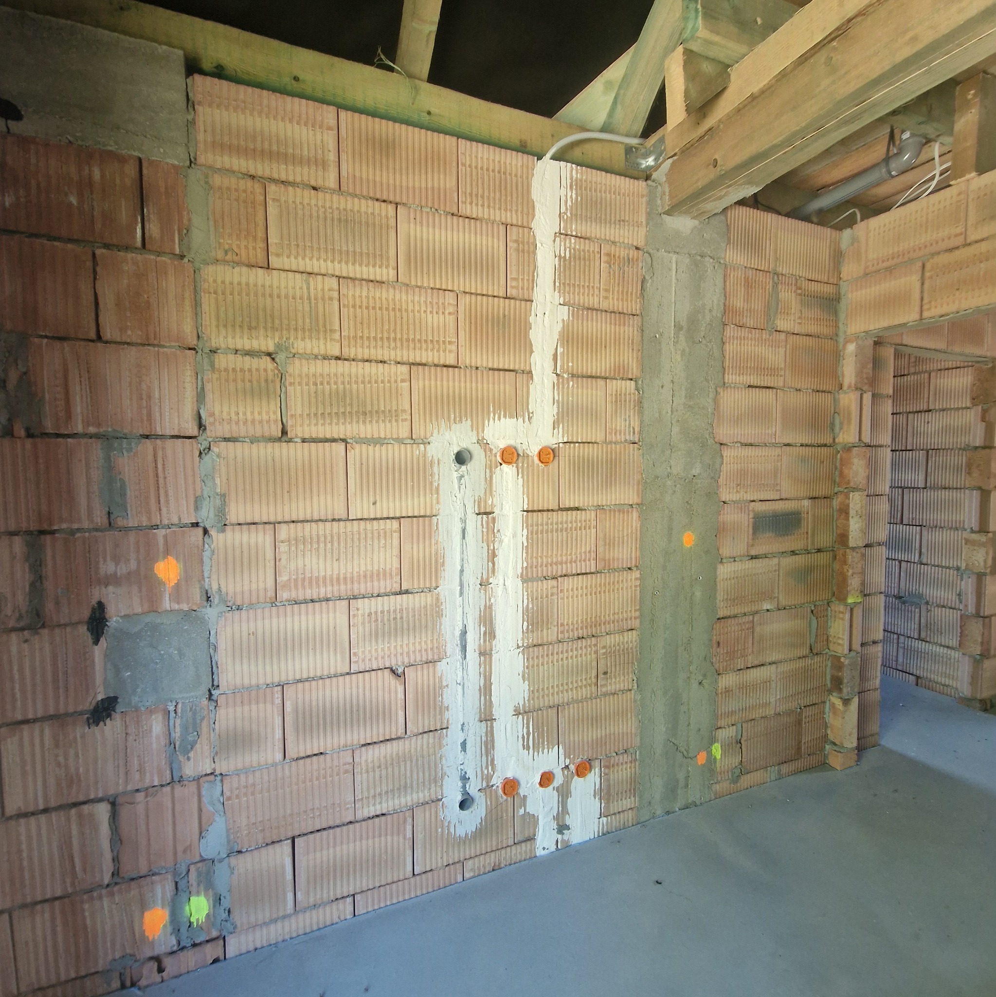Surowy mur z cegły z wyfrezowanymi bruzdami pod instalację elektryczną, zabezpieczonymi pomarańczowymi zaślepkami. Widoczne elementy konstrukcji stropu.