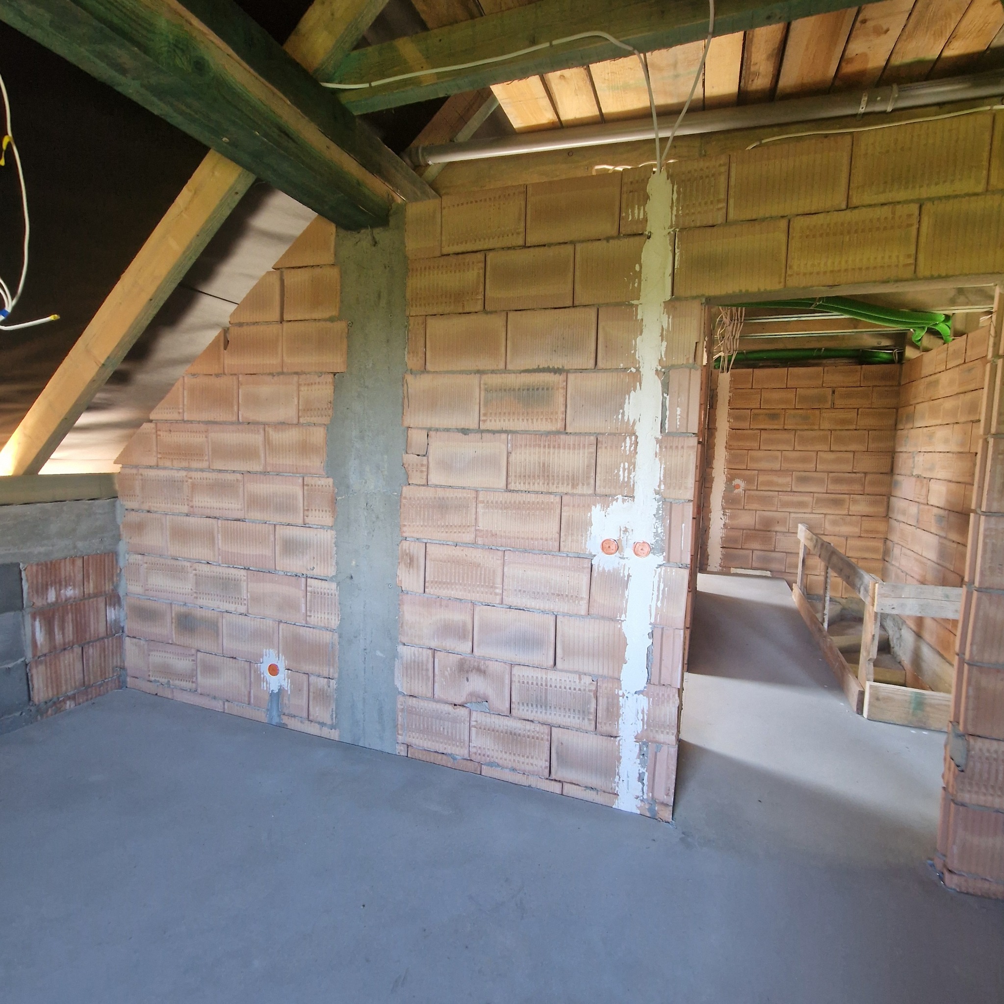 Surowy stan poddasza z ceglanymi ścianami, betonową podłogą i drewnianą konstrukcją dachu. Widoczne instalacje elektryczne i fragmenty ścian przygotowane do dalszych prac.