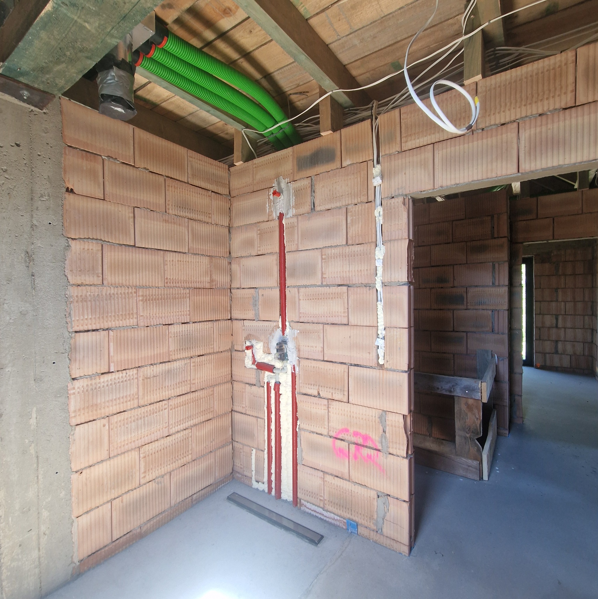 Surowa ściana z pustaków ceramicznych z instalacją wodną w czerwonych rurach, zabezpieczoną pianką montażową. Widoczne zielone rury instalacji pod sufitem i betonowy filar.
