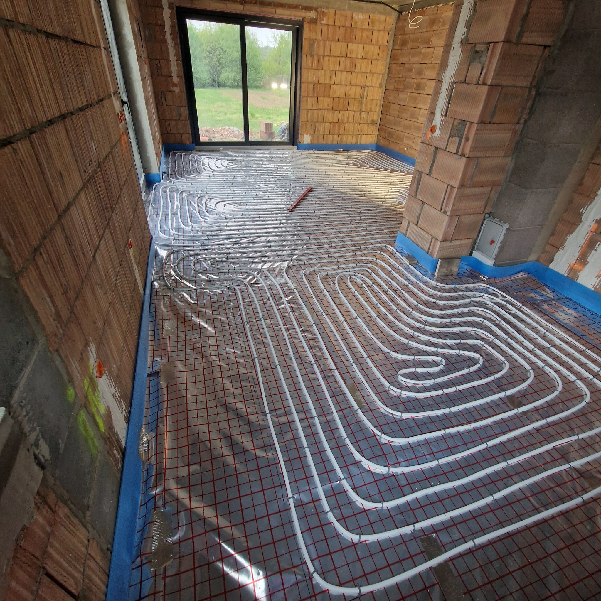 Instalacja ogrzewania podłogowego w surowym wnętrzu z ceglanymi ścianami. Białe rurki ułożone na czerwonej siatce, widoczne okno i fragment ściany z instalacją elektryczną.