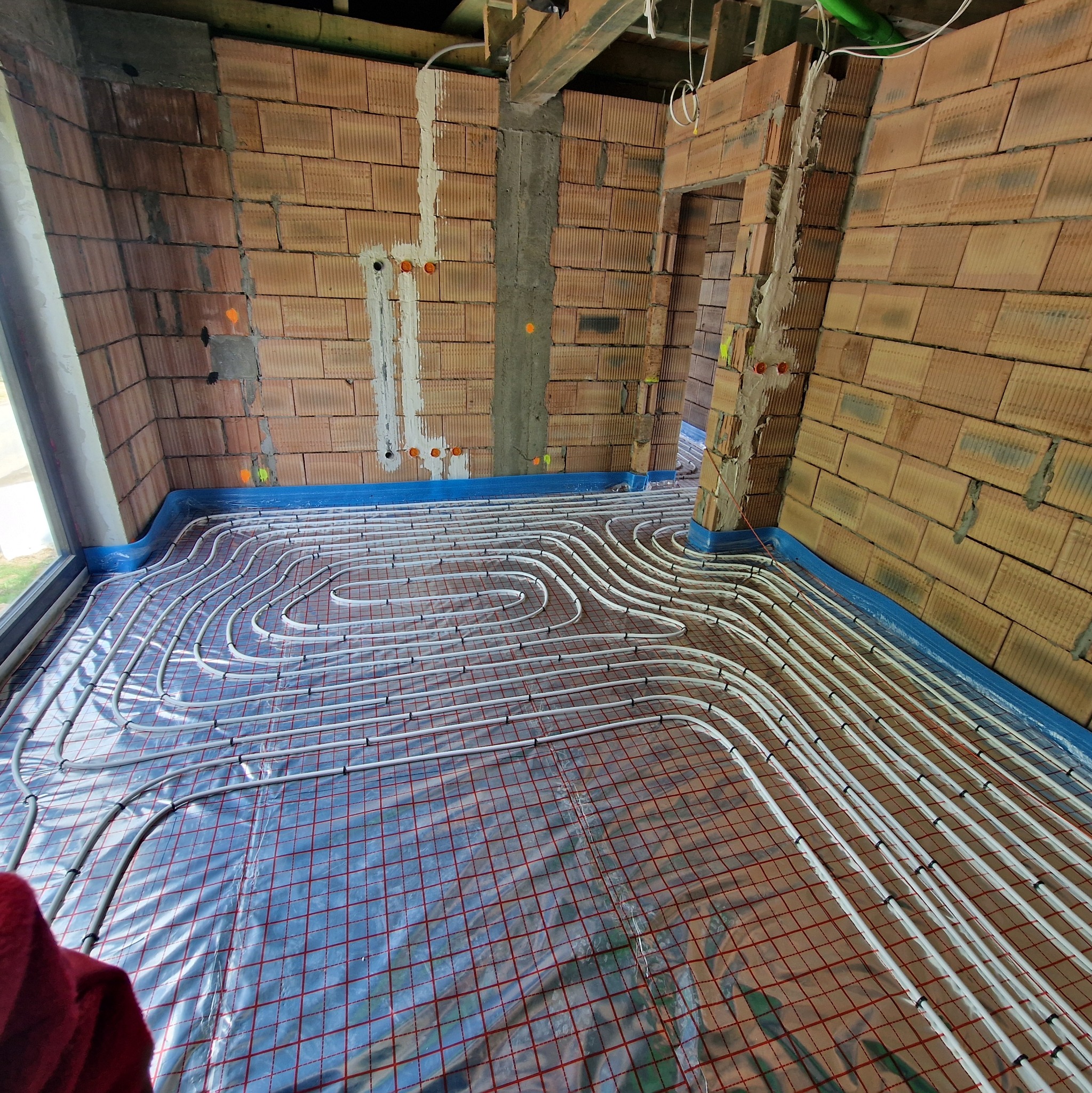 Instalacja ogrzewania podłogowego w surowym wnętrzu z ceglanymi ścianami. Białe rurki ułożone na folii z czerwoną siatką, widoczne piony instalacyjne w ścianach.