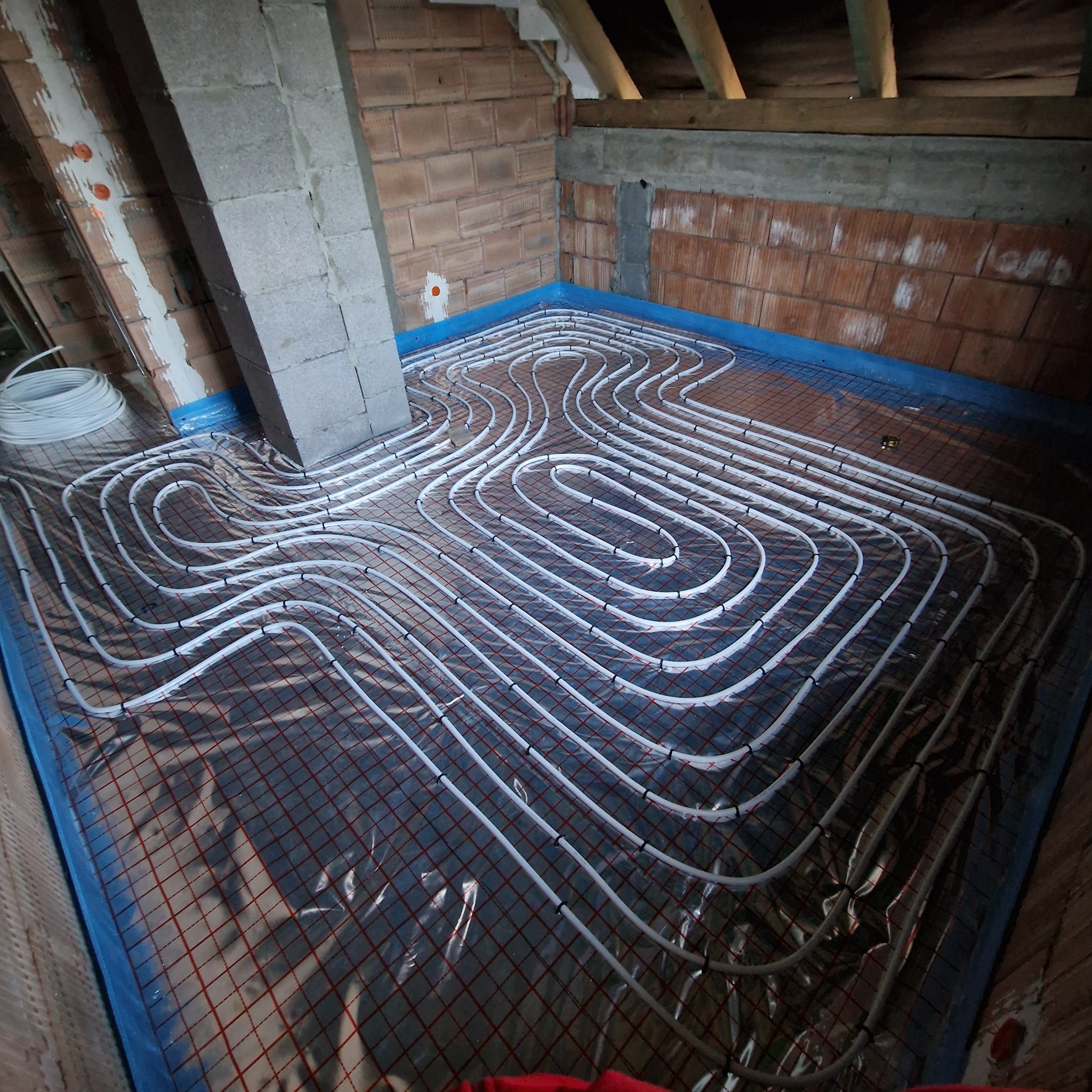 Białe rury ogrzewania podłogowego ułożone na folii z siatką w pomieszczeniu z ceglanymi ścianami i drewnianą konstrukcją dachu. Widok z góry na instalację przed wylaniem posadzki.