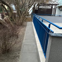 Mykhailo Mykhailiyk - Wykończony balkon z szarą podłogą z płytek i niebieską barierką. Widoczne drzewa i krzewy w tle. Ujęcie z dołu, podkreślające detale wykończenia.