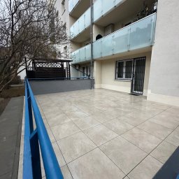 Mykhailo Mykhailiyk - Wyłożony jasnymi płytkami balkon w bloku z niebieską barierką. Widok na budynek z przeszklonymi balustradami i drzewo bez liści. Nowoczesne wykończenie.