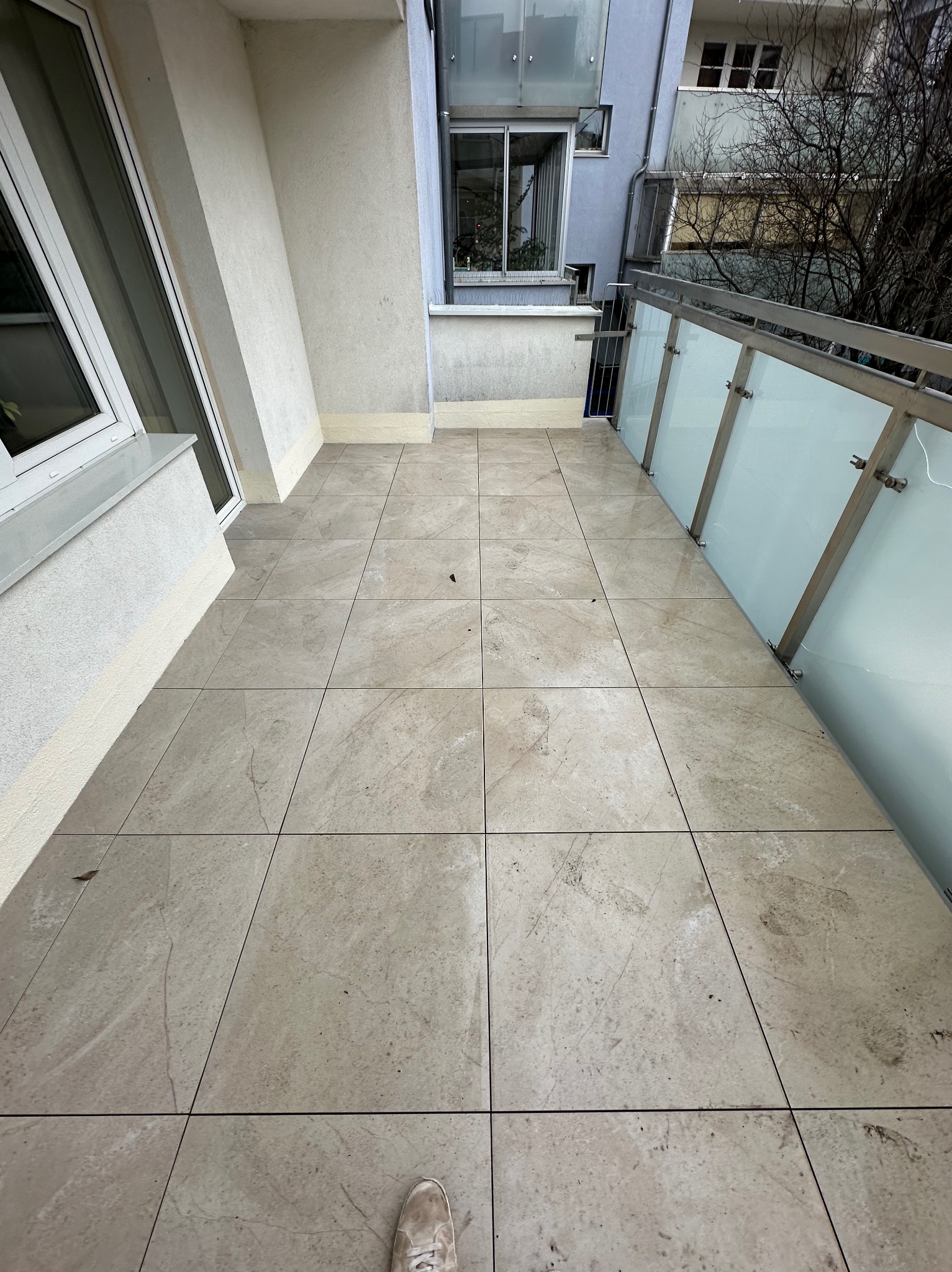 Wykończony balkon z beżowymi płytkami podłogowymi i szklaną balustradą. Widok z góry na świeżo położoną podłogę, z fragmentem buta w rogu kadru.