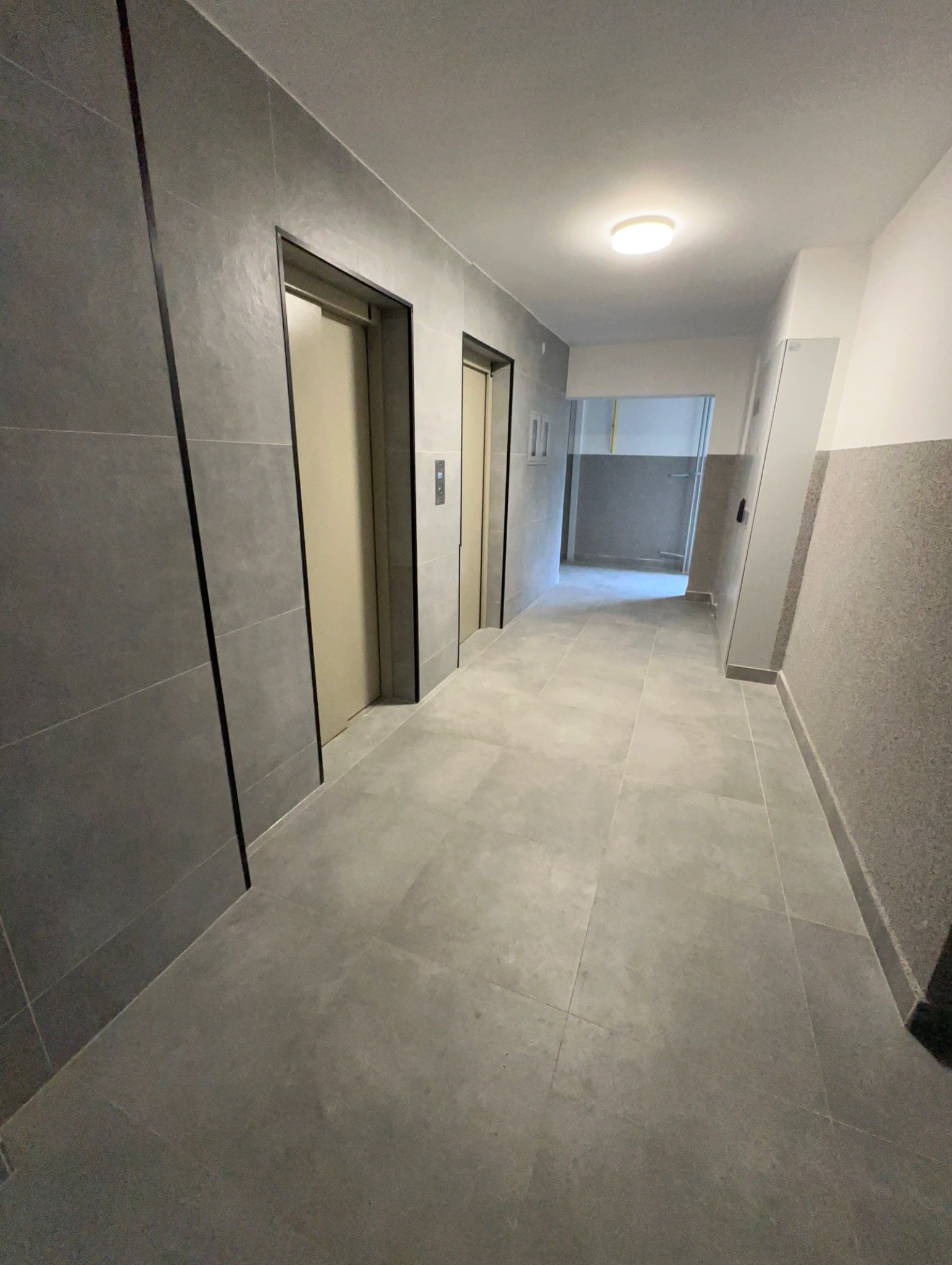 Wykończony korytarz w bloku z szarymi płytkami na podłodze i ścianach. Dwa wejścia do windy po lewej stronie. Widok w głąb korytarza.