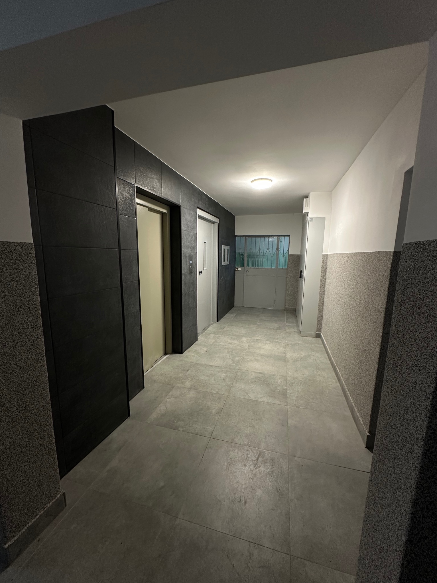 Wyremontowany korytarz z szarymi płytkami podłogowymi i ciemną okładziną ścienną przy windzie. Widoczne drzwi i okno na końcu korytarza.