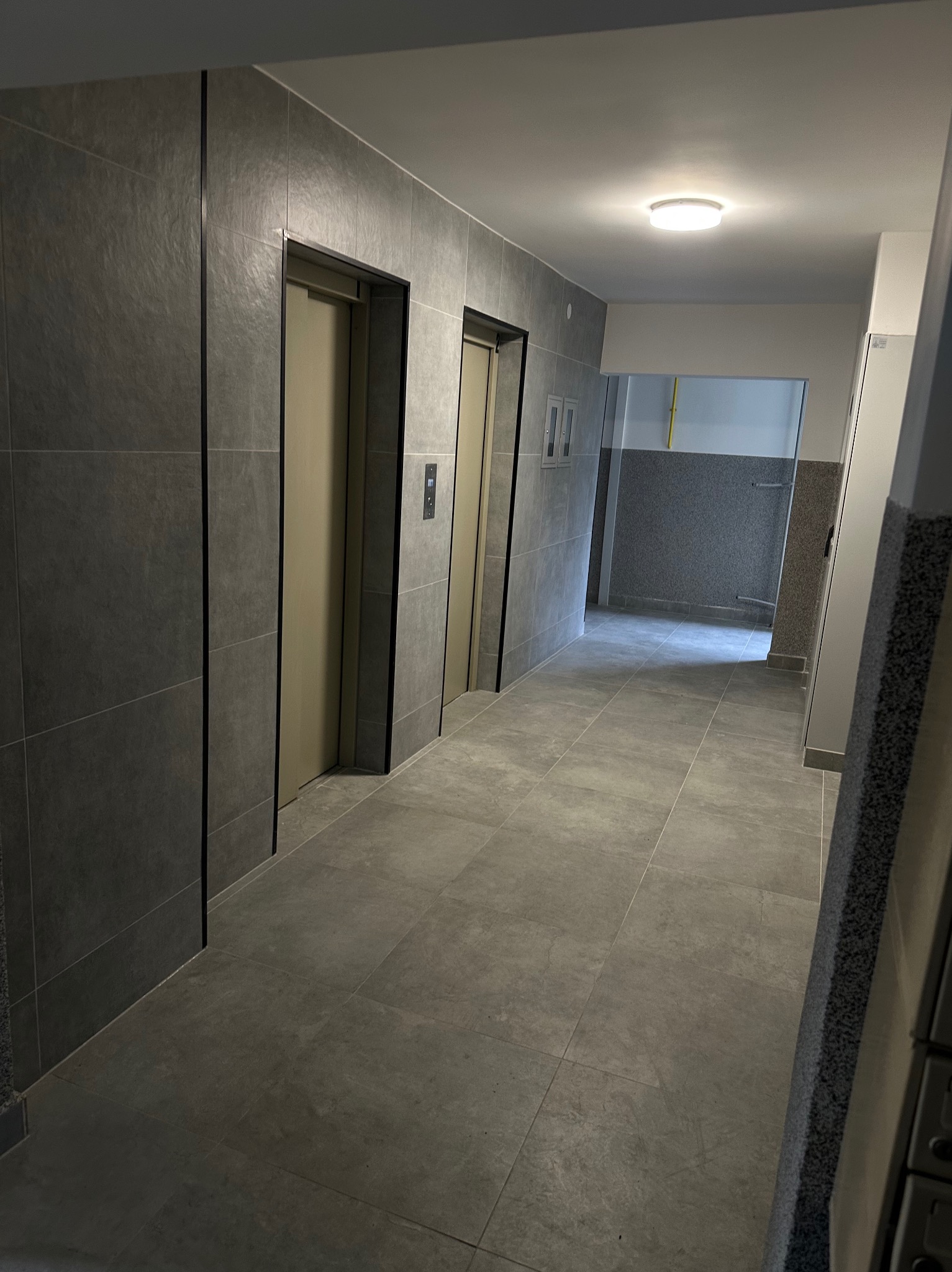 Korytarz w budynku wielorodzinnym po remoncie. Szare płytki na podłodze i ścianach, dwie windy z beżowymi drzwiami. Widoczne detale wykończenia.