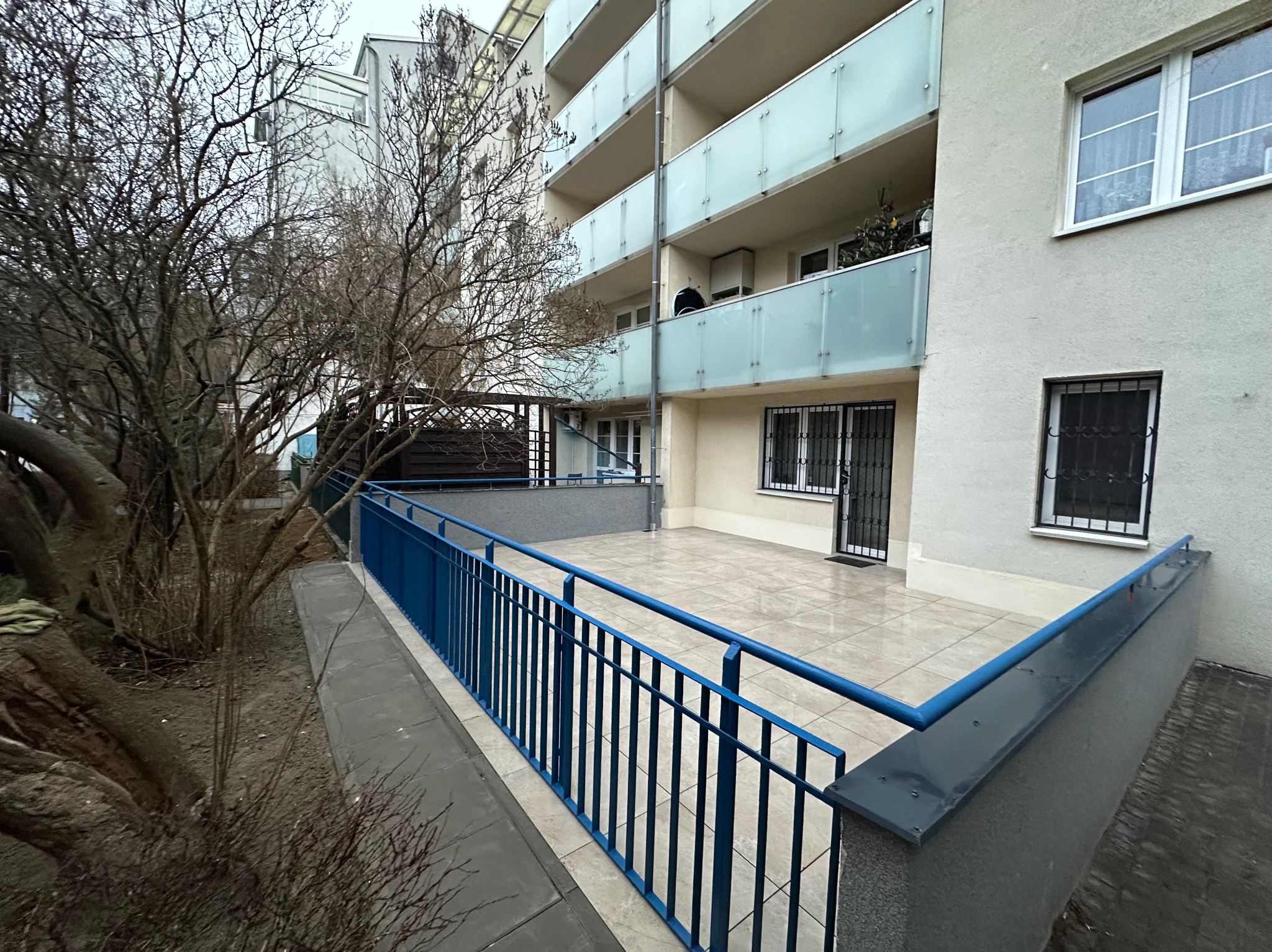 Wyremontowany balkon z niebieską barierką i jasnymi płytkami. Widok z dołu na budynek z balkonami. Drzewa bez liści po lewej stronie kadru. Dobre oświetlenie.