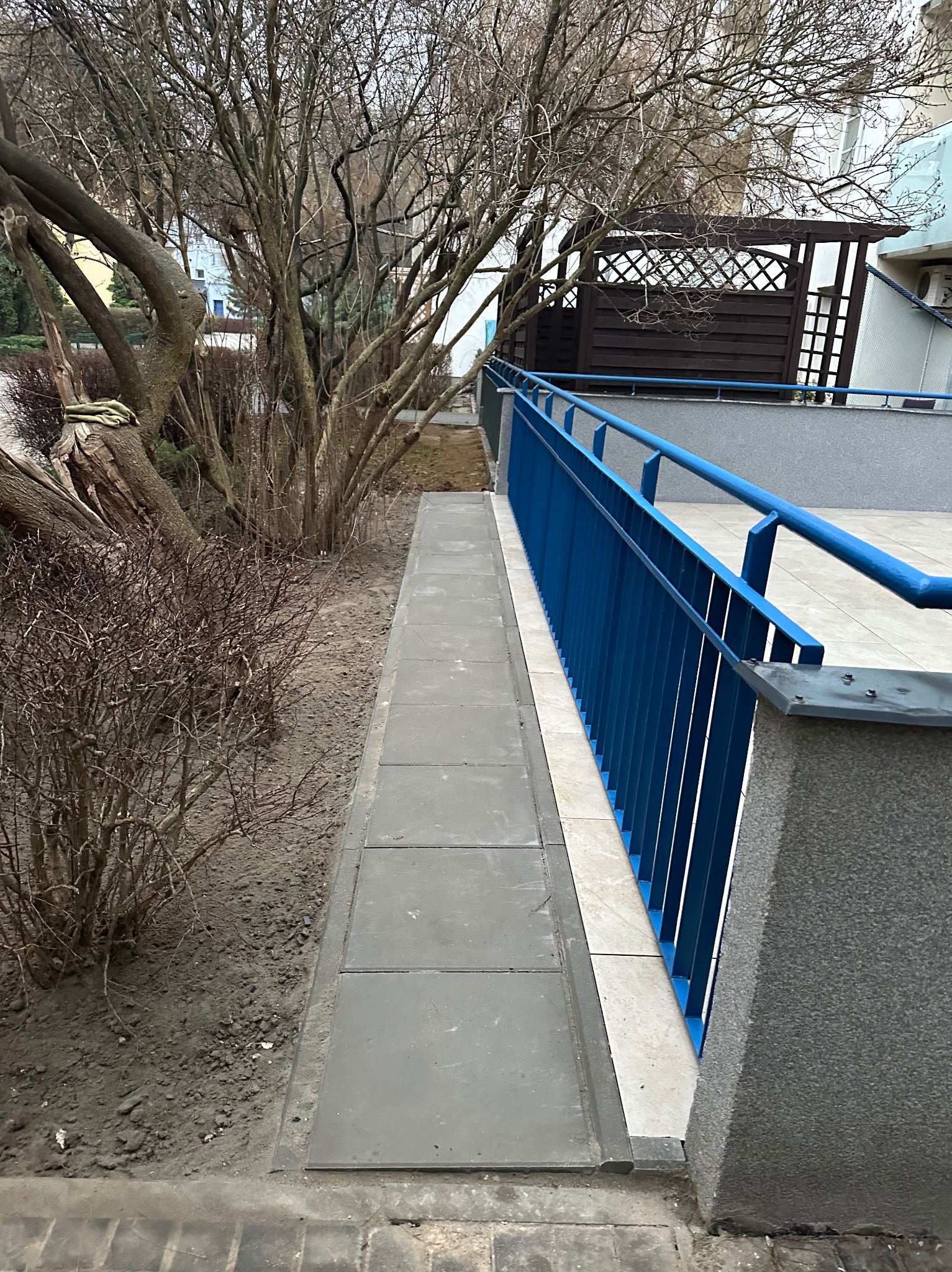 Wykończony balkon z szarą podłogą z płytek i niebieską barierką. Widoczne drzewa i krzewy w tle. Ujęcie z dołu, podkreślające detale wykończenia.