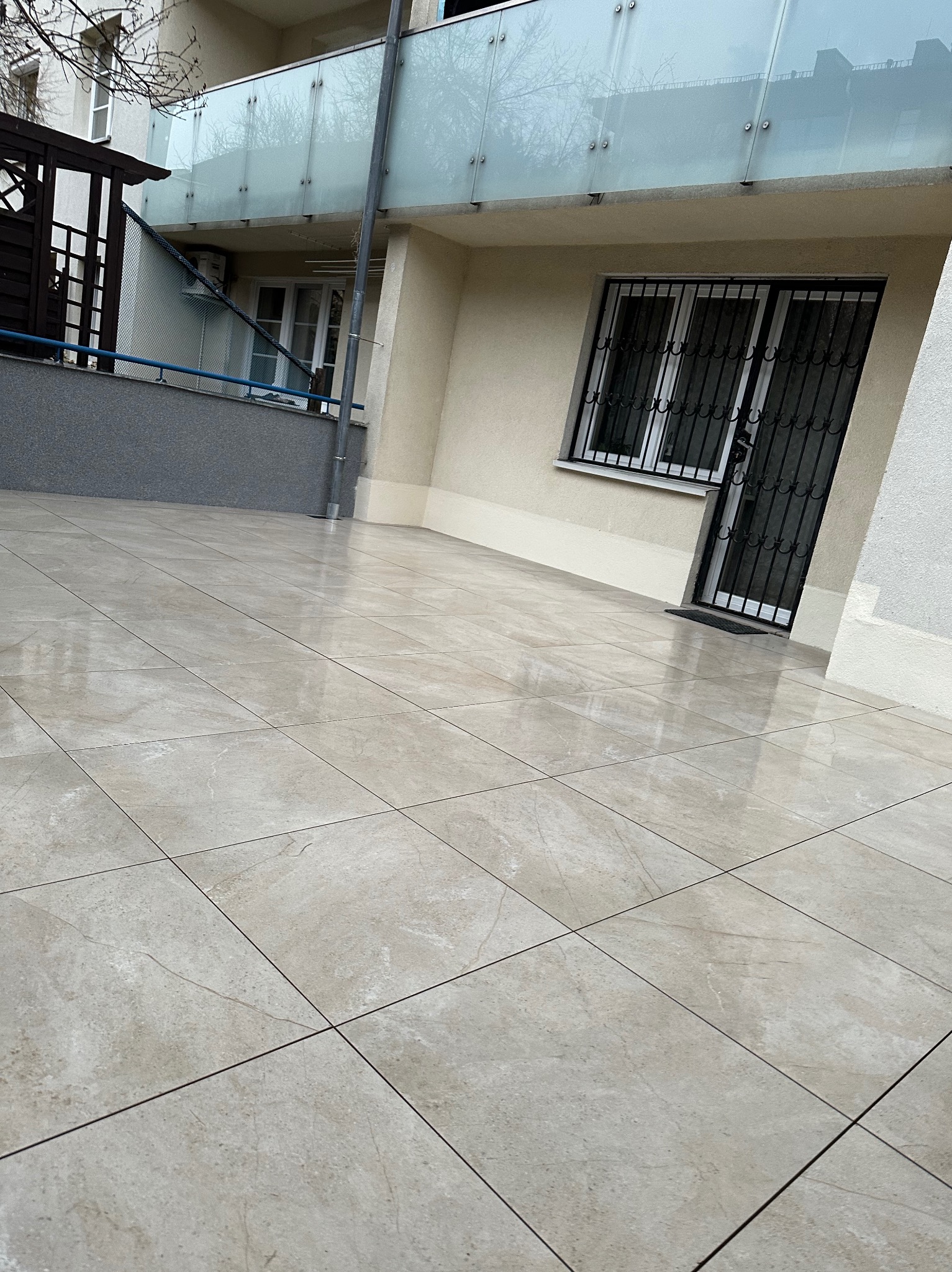 Jasne, błyszczące płytki na balkonie z przeszkloną balustradą i oknem z kratą. Ułożenie płytek w karo, widok z dołu, lekki kąt.