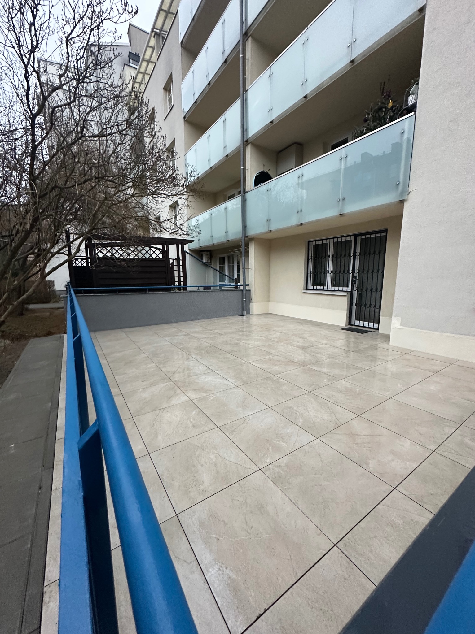 Wyłożony jasnymi płytkami balkon w bloku z niebieską barierką. Widok na budynek z przeszklonymi balustradami i drzewo bez liści. Nowoczesne wykończenie.