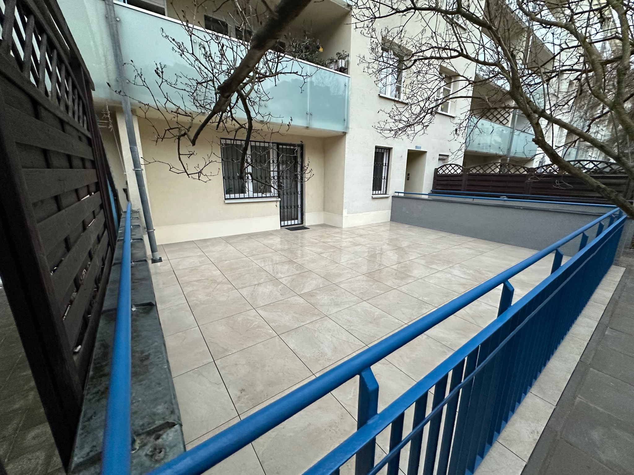 Wyłożony jasnymi płytkami taras z niebieską barierką, widok na budynek z balkonami. Minimalistyczny design, świeżo po remoncie, czysta przestrzeń.