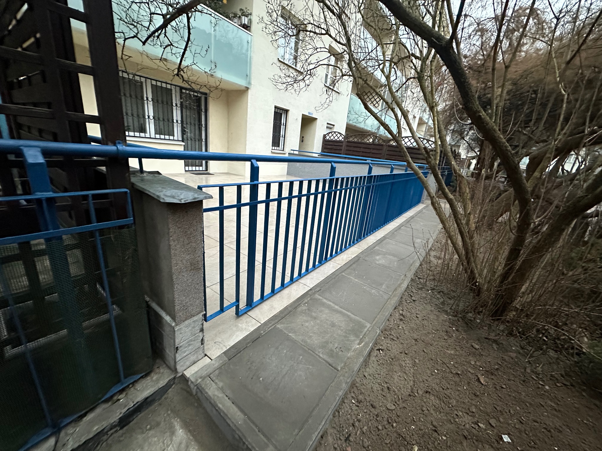 Niebieska balustrada balkonu z widokiem na budynek mieszkalny i drzewa bez liści. Ujęcie z dołu, podkreślające perspektywę i detale wykończenia.