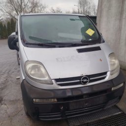 Kamil Niedrygoś - Srebrny bus Opel Vivaro z żółtą kartką za szybą, zaparkowany na zewnątrz, widok z przodu, lekko zanieczyszczony, zarys drzew na drugim planie.