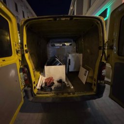 Kamil Niedrygoś - Żółty bus z otwartymi drzwiami, załadowany meblami i kartonami, gotowy do transportu na tle nocnego krajobrazu miejskiego.