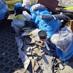 Kamil Niedrygoś - Sterta gruzu i worków z odpadami budowlanymi na kostce brukowej, po remoncie. Widoczne fragmenty płytek, wiadro z farbą i inne pozostałości. Słoneczny dzień.