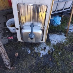 Kamil Niedrygoś - Używana szafka z metalowym zlewem, wystawiona na zewnątrz na trawie z resztkami śniegu. Widoczny biały pojemnik i niebieski worek. Ujęcie z bliska, z dołu.