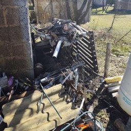 Kamil Niedrygoś - Sterta różnorodnych odpadów i złomu w zaniedbanym otoczeniu, prawdopodobnie przed opróżnianiem. Widoczne elementy konstrukcyjne budynku i zieleń w tle.