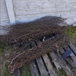 Kamil Niedrygoś - Stara, zardzewiała siatka ogrodzeniowa i drewniane deski leżące na trawie przy betonowym murze. Elementy po demontażu, przygotowane do wywozu.