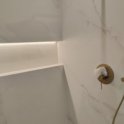 BUD PROFIL - Wykończona wnęka z oświetleniem LED i bateria prysznicowa w kolorze złota na marmurowej ścianie w łazience we Wrocławiu. Minimalistyczny design.