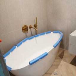 BUD PROFIL - Nowoczesna, wolnostojąca wanna owalna z złotą armaturą, zabezpieczona niebieską pianką. Jasne płytki na ścianach i drewnopodobna podłoga. Częściowo widoczna szafka.