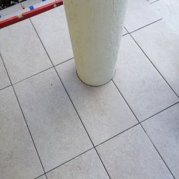 BUD PROFIL - Ułożone jasne płytki podłogowe wokół donicy, widoczna poziomica i worek z fugą. Detal wykończenia tarasu lub balkonu po remoncie, z bliska.