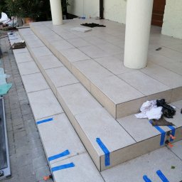 BUD PROFIL - Kładzenie jasnych płytek na schodach wejściowych z kolumnami. Widoczne narzędzia i taśmy mocujące. Częściowo ukończony etap prac glazurniczych na zewnątrz budynku.