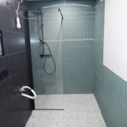 BUD PROFIL - Wykończona kabina prysznicowa z ciemnymi i jasnymi płytkami. Widoczna armatura, podłączenie elektryczne i fragment podłogi z mozaiki. Minimalistyczny design.