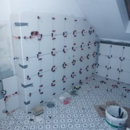 BUD PROFIL - Białe płytki z systemem poziomowania na ścianach i podłodze w łazience na poddaszu. Widoczne narzędzia i wiadro z zaprawą. Etap prac glazurniczych.