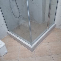 BUD PROFIL - Nowoczesna kabina prysznicowa z marmurowym wykończeniem, widok z dołu, ukazujący detale płytek i brodzika. Chromowana armatura i szklane drzwi.