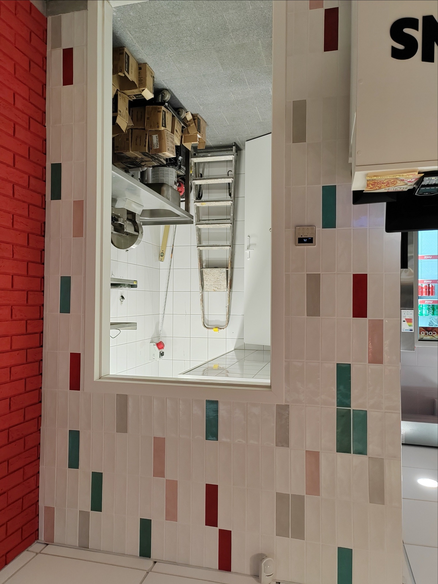 Białe, wąskie płytki z kolorowymi akcentami na ścianie, widok na zaplecze z metalowymi elementami i drabiną. Czerwona ściana z cegły w lewym rogu.