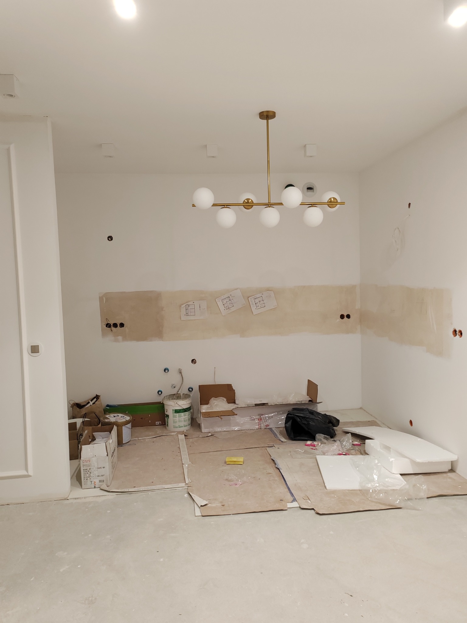 Wnętrze w trakcie prac wykończeniowych: ściany po szpachlowaniu, widoczne instalacje elektryczne i hydrauliczne, podłoga zabezpieczona kartonem, nowoczesna lampa sufitowa.