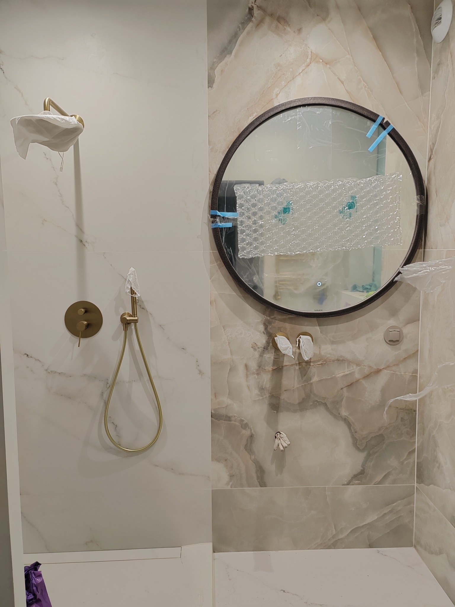 Wykończona łazienka z marmurowymi płytkami, złotą armaturą i okrągłym lustrem, jeszcze zabezpieczonym folią ochronną. Nowoczesny design.
