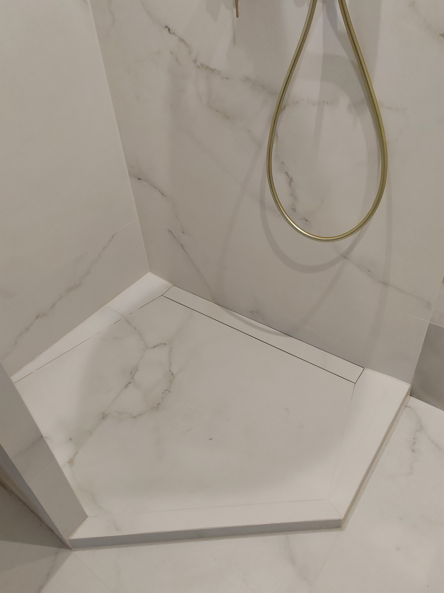 Wykończona kabina prysznicowa z białymi płytkami imitującymi marmur i złotym uchwytem na słuchawkę. Widok z góry, detale wykończenia.