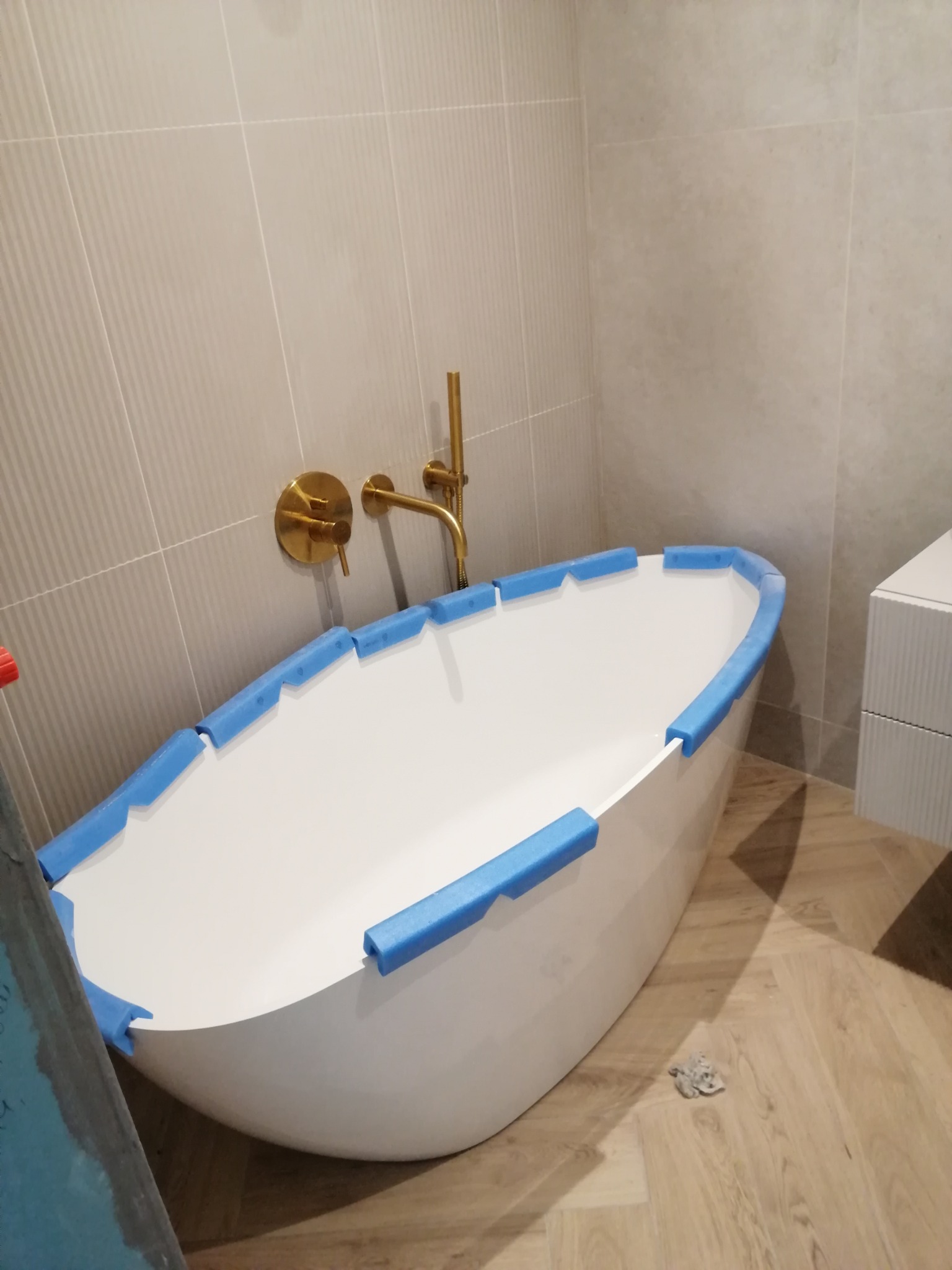 Nowoczesna, wolnostojąca wanna owalna z złotą armaturą, zabezpieczona niebieską pianką. Jasne płytki na ścianach i drewnopodobna podłoga. Częściowo widoczna szafka.