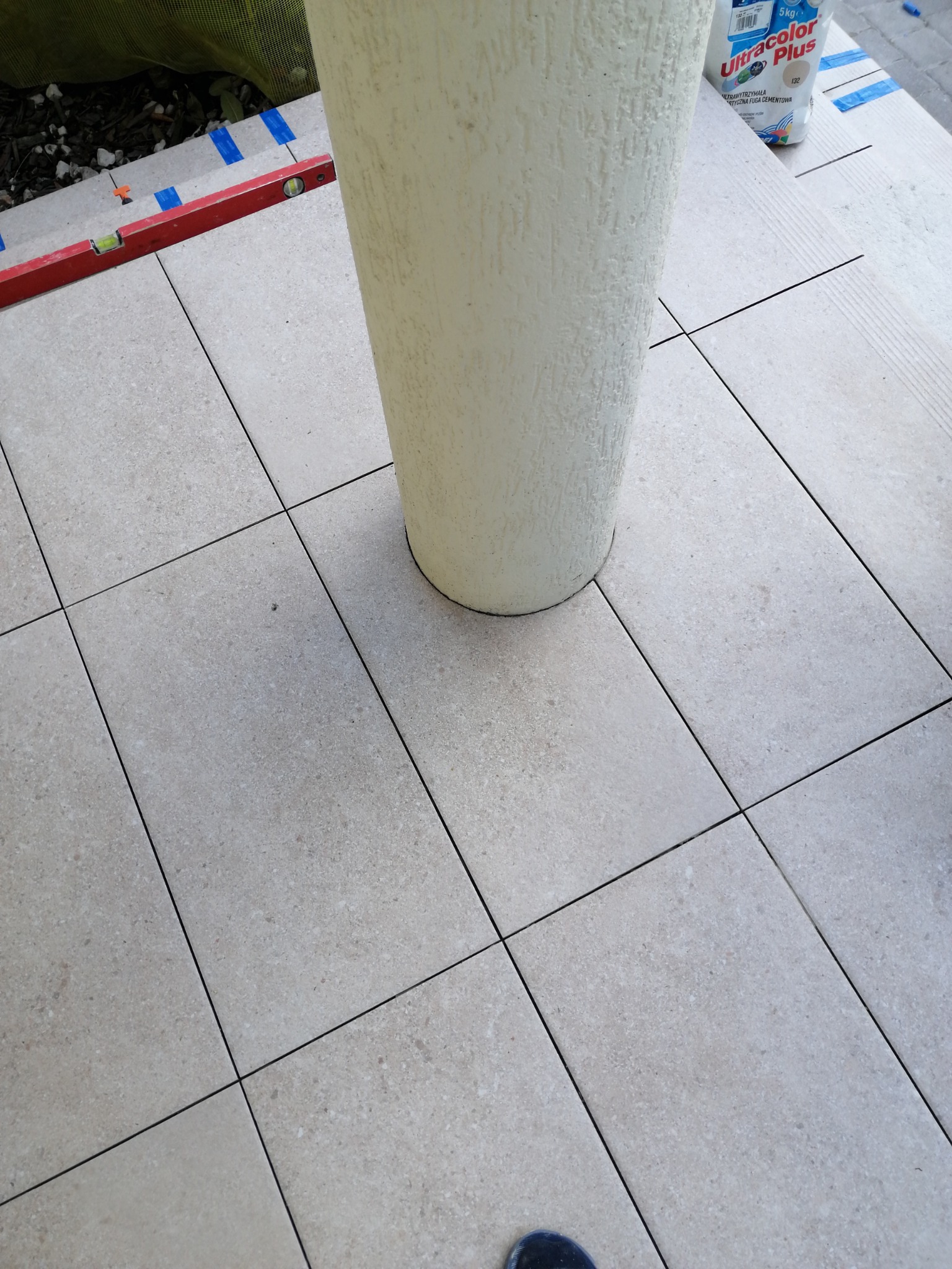 Ułożone jasne płytki podłogowe wokół donicy, widoczna poziomica i worek z fugą. Detal wykończenia tarasu lub balkonu po remoncie, z bliska.