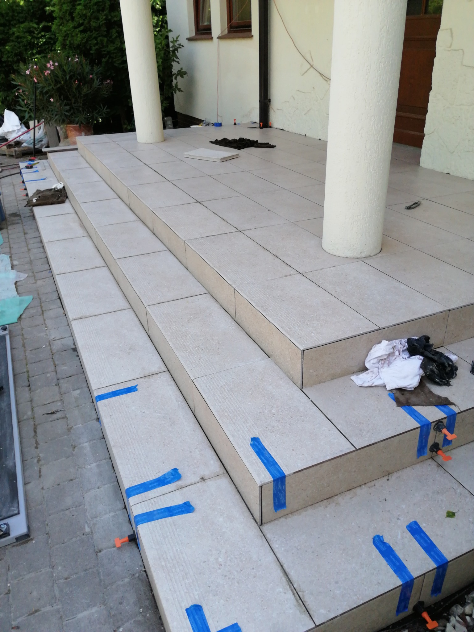 Kładzenie jasnych płytek na schodach wejściowych z kolumnami. Widoczne narzędzia i taśmy mocujące. Częściowo ukończony etap prac glazurniczych na zewnątrz budynku.