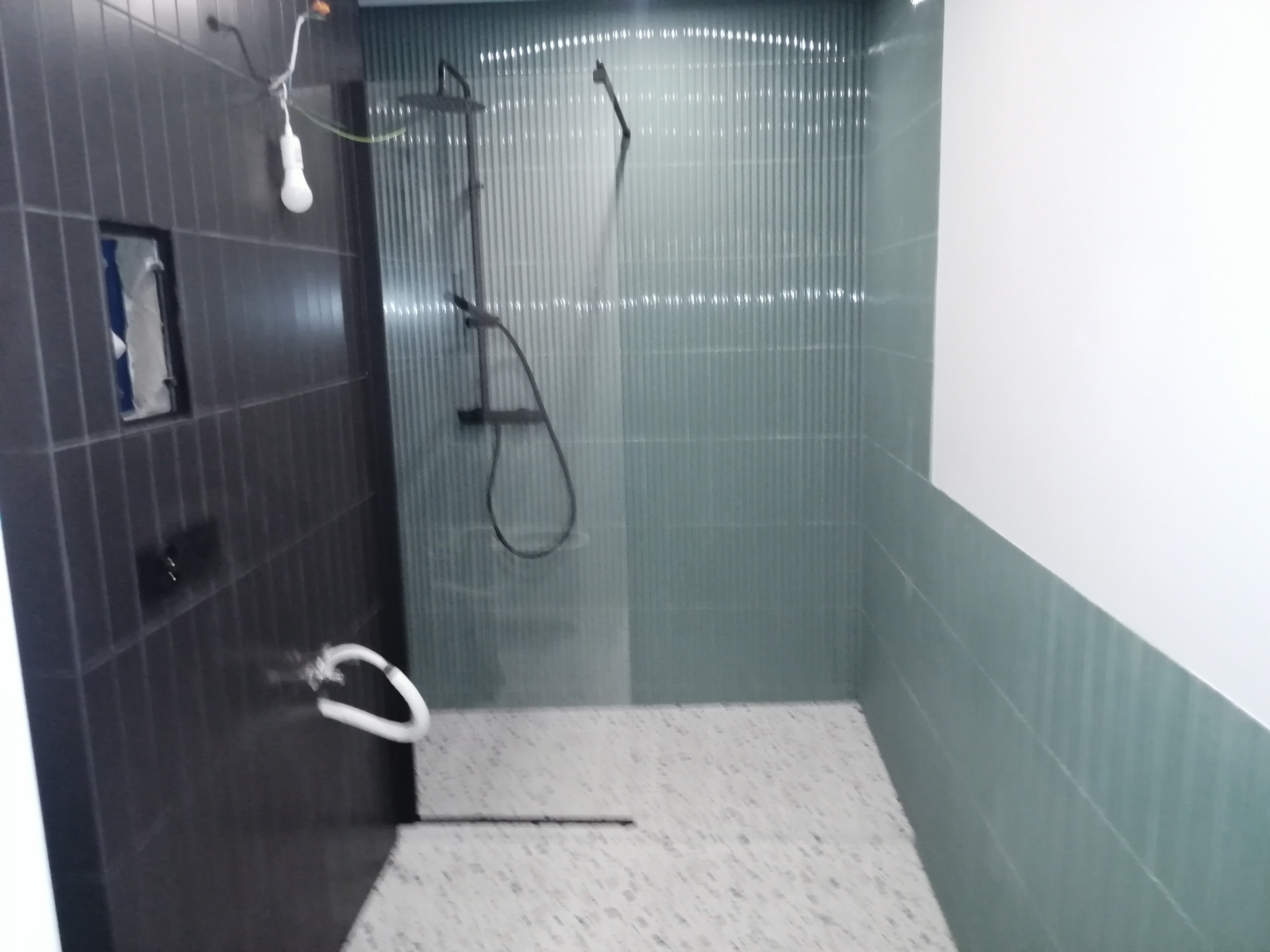 Wykończona kabina prysznicowa z ciemnymi i jasnymi płytkami. Widoczna armatura, podłączenie elektryczne i fragment podłogi z mozaiki. Minimalistyczny design.