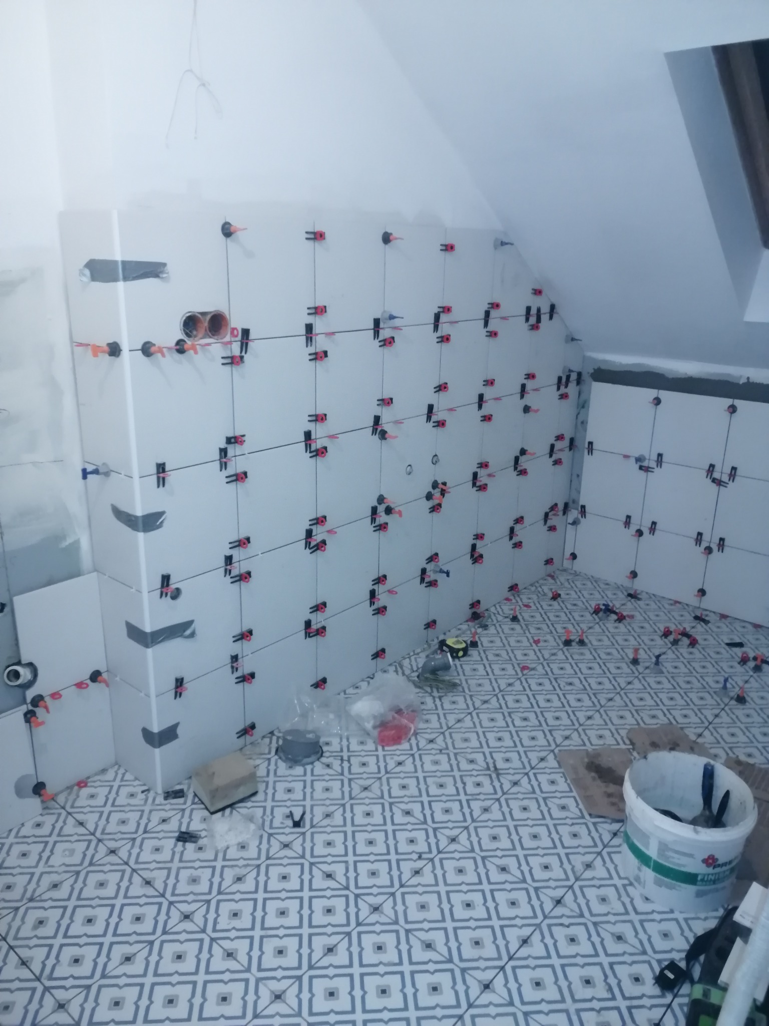 Białe płytki z systemem poziomowania na ścianach i podłodze w łazience na poddaszu. Widoczne narzędzia i wiadro z zaprawą. Etap prac glazurniczych.